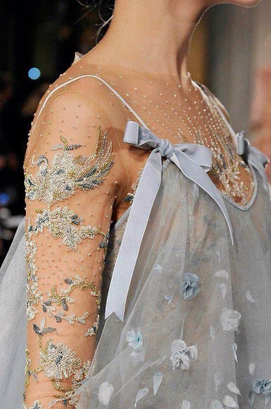 a closeup shot of a woman wearing a beautiful, ethereal gown. the dress is made of sheer tulle, and the sleeves are adorned with intricate gold and silver embroidery. the dress is cinched at the waist with a blue velvet ribbon. the model is standing with her arms at her side and she is looking slightly down at the ground. the dress is so delicate and intricate that it looks like a work of art.