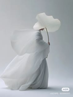 a woman in a flowing white dress stands gracefully, holding a delicate white leaf in her hand. the image is minimal and serene, with a soft focus and a pale white background. the woman's pose and the delicate leaf create a sense of elegance and grace. the overall effect is one of ethereal beauty and tranquility.