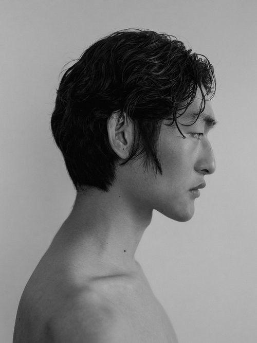 a black and white portrait of a young man looking away from the camera. his hair is slicked back and he is wearing a simple white shirt. the background is a plain white wall, and the lighting is soft and natural. the man's expression is serious and intense.