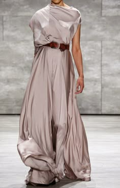 a fashion model in a long, gray gown with a brown belt poses on a runway. the dress is flowy and elegant, with a dramatic drape. the model has her hair styled in a simple updo and her makeup is minimal. the background is a plain grey wall, highlighting the dress and model's features.  the lighting is soft and diffused, creating a dreamy atmosphere.
