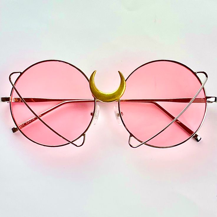 a pair of pink round framed sunglasses with a gold crescent moon connecting the lenses is displayed against a white background.