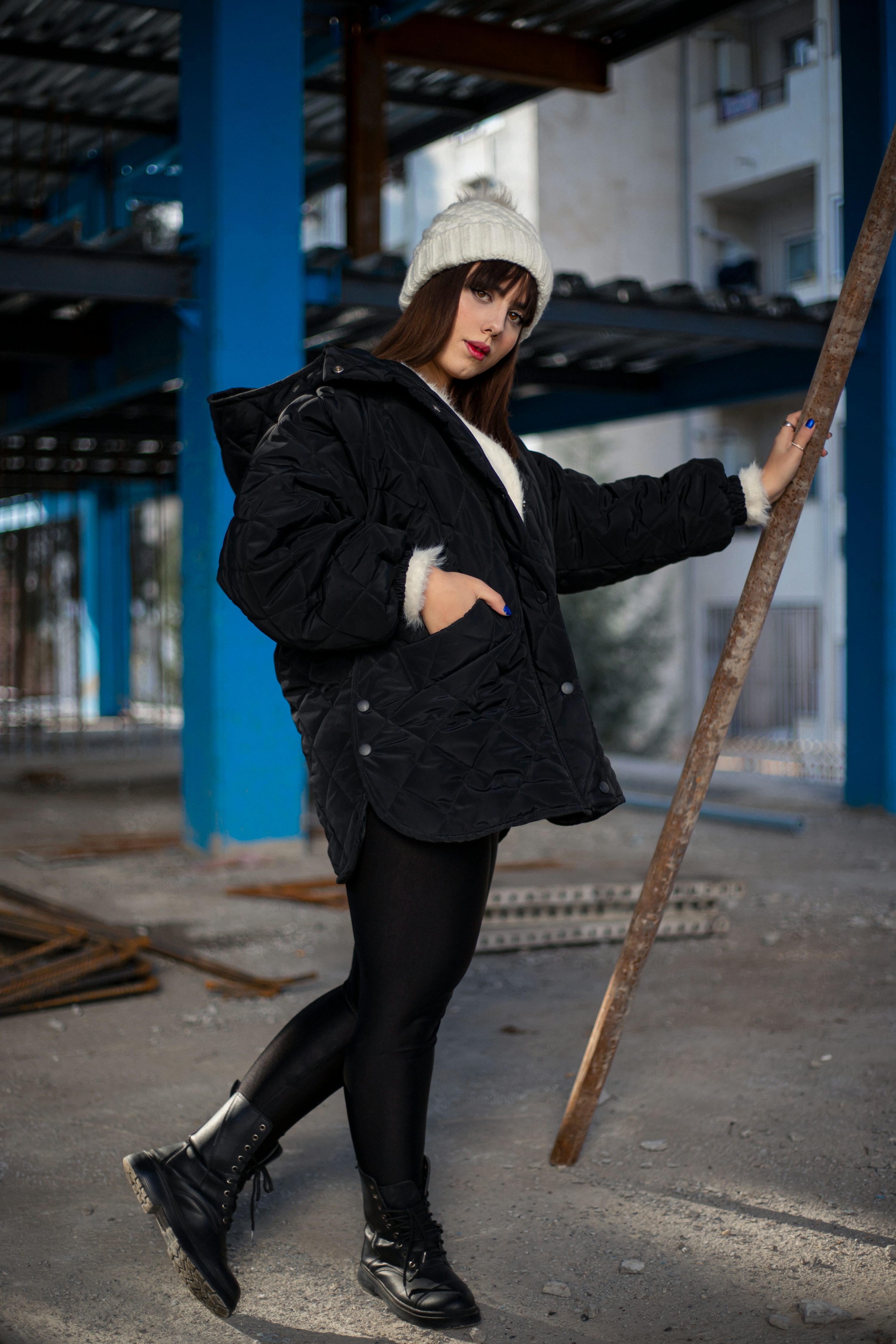 a young woman in a black quilted jacket and white beanie leans against a wooden pole on a construction site. she is wearing black leggings and combat boots, and her overall style is urban and trendy. the photo has a cool and confident vibe.