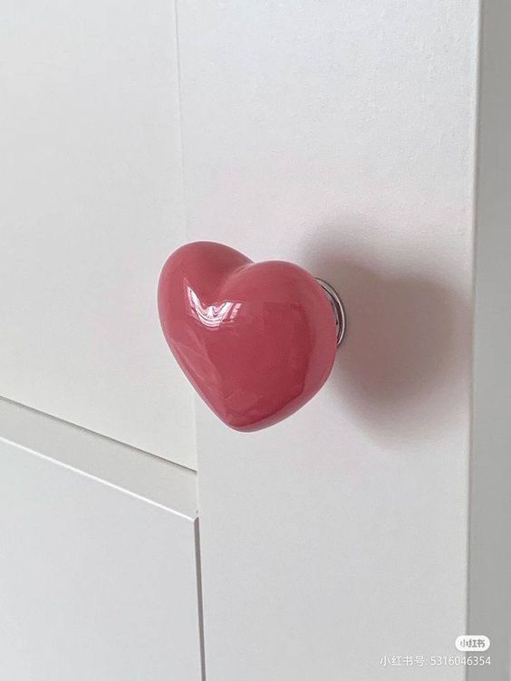 a closeup of a pink heart shaped door knob on a white cabinet door. the door knob is made of ceramic and has a glossy finish. the background is a white cabinet door.  the door knob is a unique and stylish way to add a touch of love to any room.