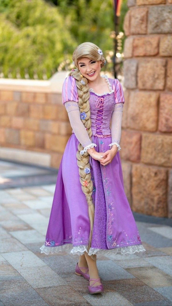 a young woman dressed in a rapunzel costume smiles at the camera.  she's wearing a long purple dress with embroidered details and a lace trim, and her long blonde braid is adorned with small flowers. she's standing on a paved walkway next to a stone wall, possibly in a theme park setting.  she appears happy and content.