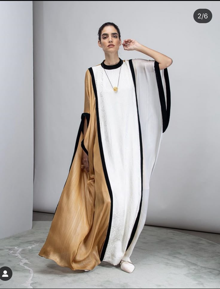 a woman is standing in a studio, posing in an elegant, asymmetrical gown. the gown is longsleeved and features a combination of gold, white, and black trim. she is wearing a necklace and the photograph is a fulllength studio shot against a neutral background.