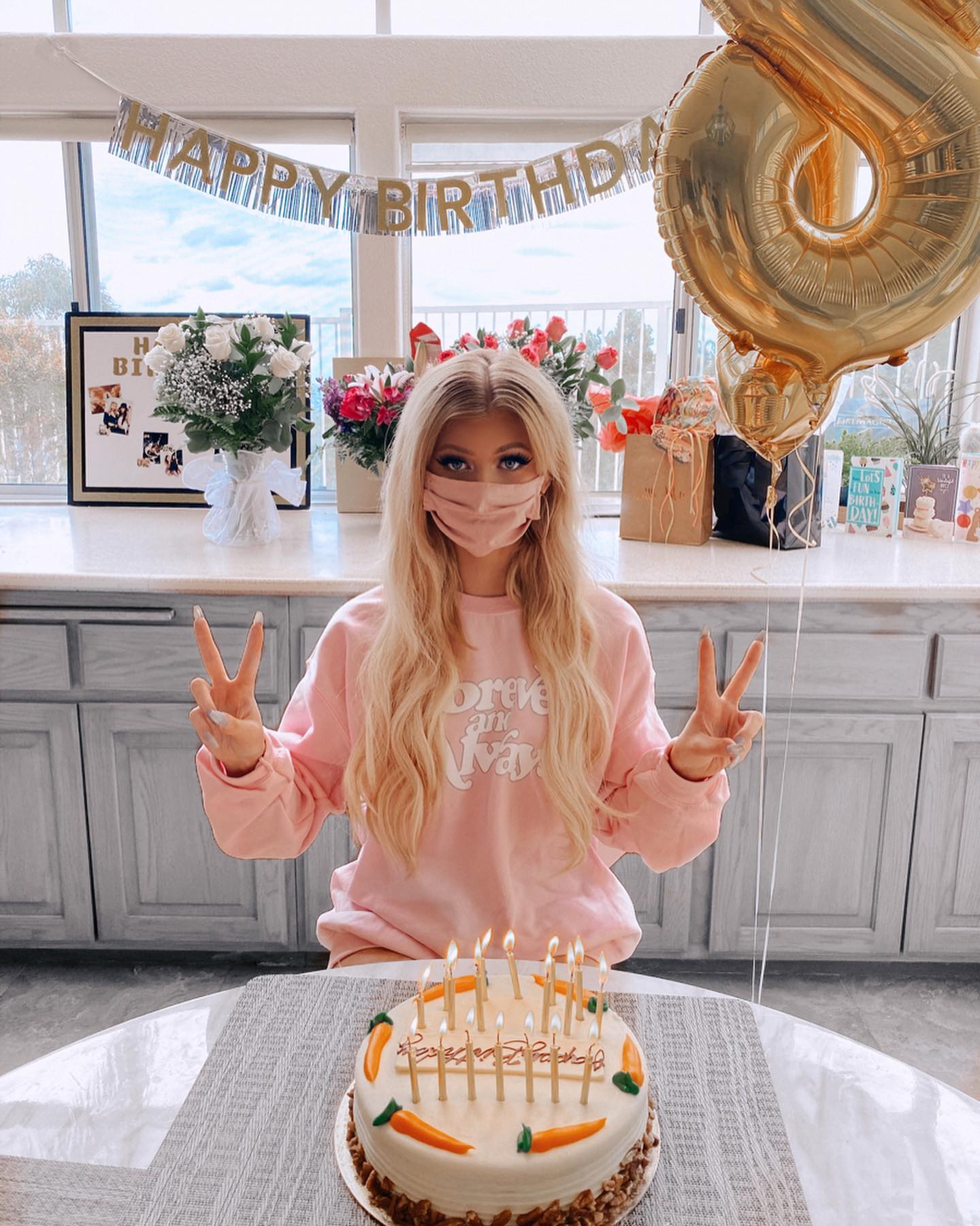 a woman wearing a pink sweater and face mask is sitting in front of a birthday cake with candles. she is making the peace sign with both hands. behind her is a window with a happy birthday banner, flowers, gifts, and a balloon in the shape of the number 18. the cake is decorated with carrots.