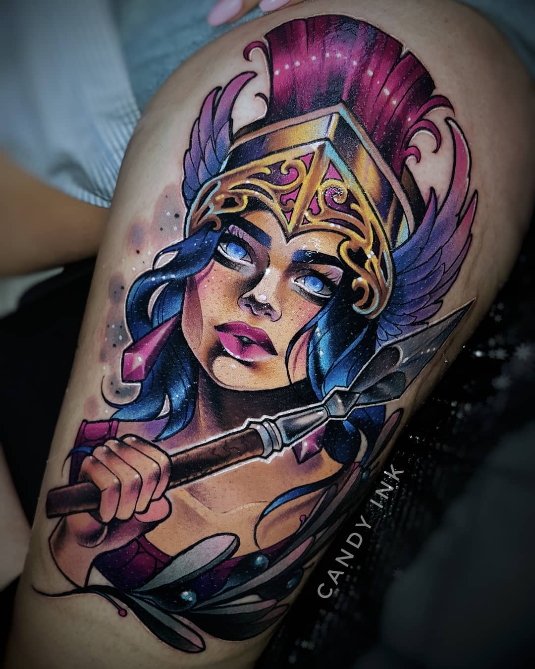 a closeup photograph showcasing a vibrant and detailed tattoo of a female warrior, possibly inspired by greek mythology, on someone's upper thigh. the warrior, depicted with striking blue eyes, holds a spear and wears an ornate helmet, suggesting strength and power. the tattoo artist's signature, "candy ink," is subtly incorporated into the design.  the image is welllit, likely taken indoors, and offers a clear, frontal perspective of the artwork.