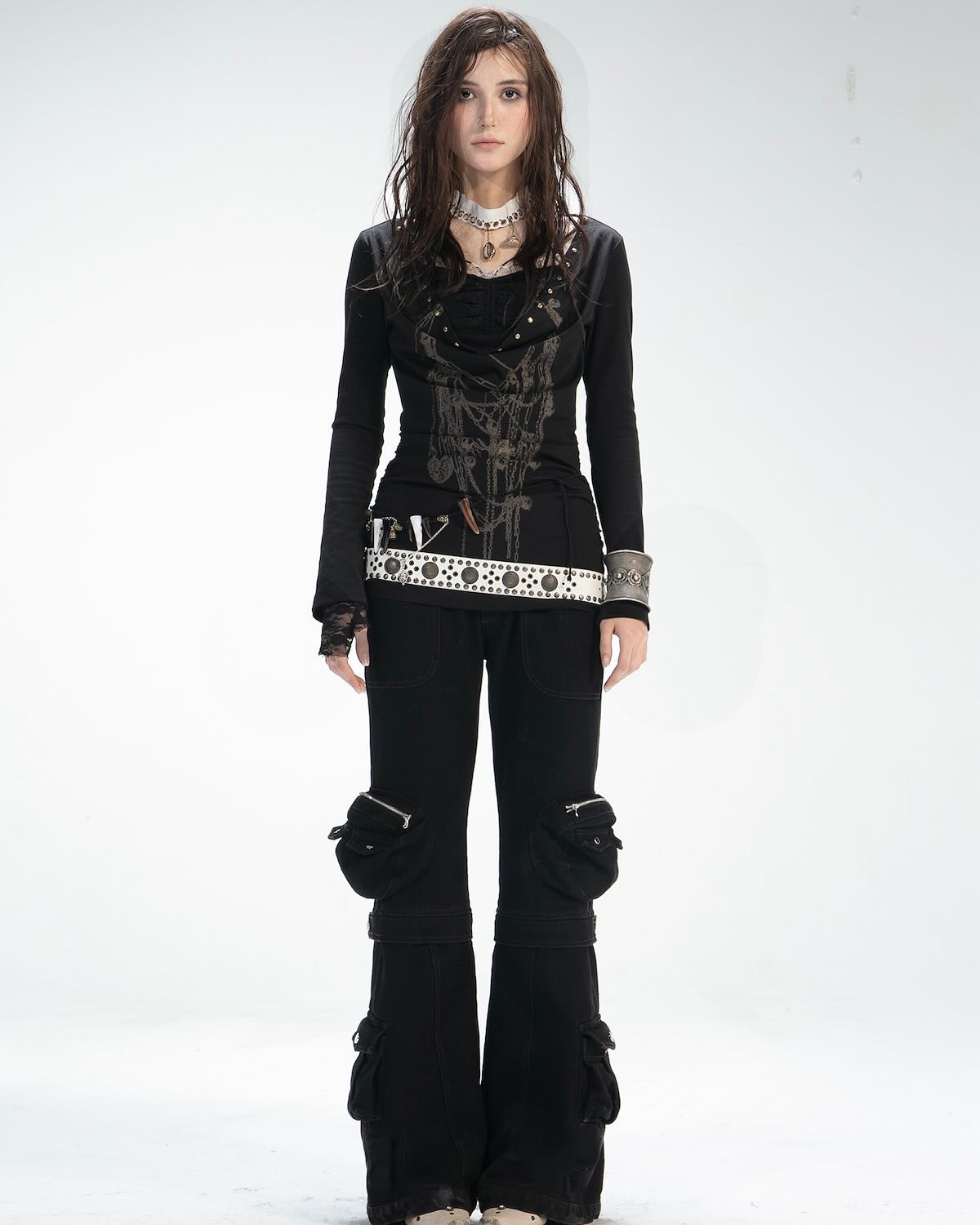 a fullbody studio shot of a young woman model posing against a white background. she is wearing a black longsleeved shirt with detailed embellishments, a white studded belt, black cargo pants with multiple pockets, and black boots. her hair is dark brown and loose. she is wearing a necklace and bracelets as accessories. the overall style is a mix of grunge, gothic, and punk elements.