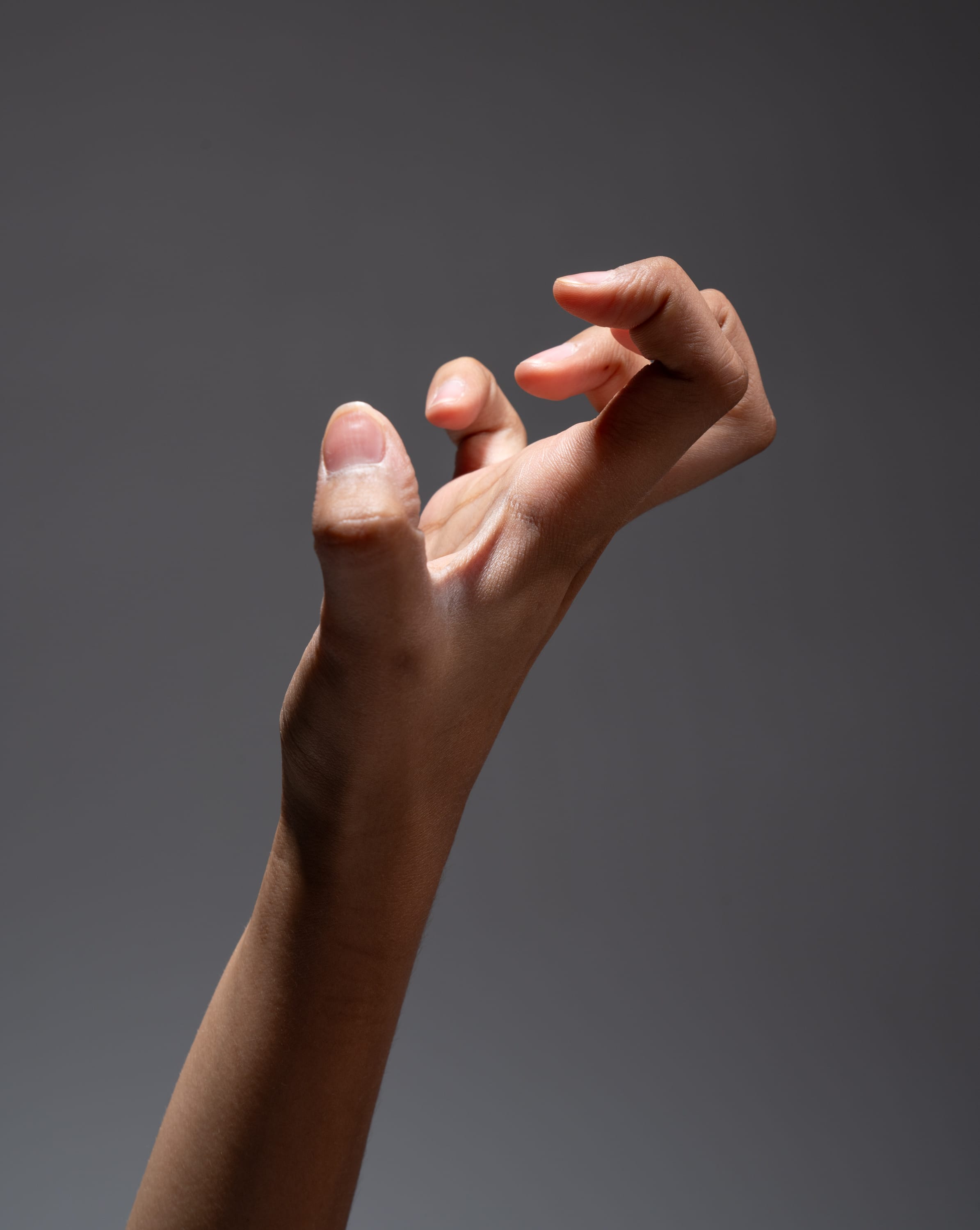 a hand is reaching upwards with the fingers slightly curled, resembling a claw. the background is a neutral gray, and the hand is lit by studio lighting, creating highlights on the skin.