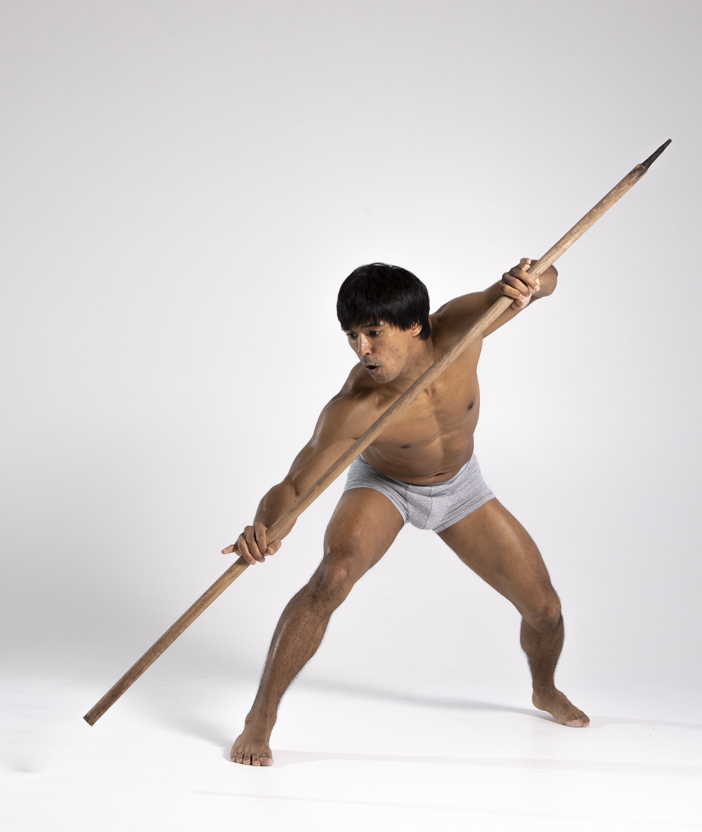 a barechested, muscular man in a gray loincloth holds a spear diagonally across his body in a fighting stance. he has dark hair and brown skin, and is barefoot. the background is white.