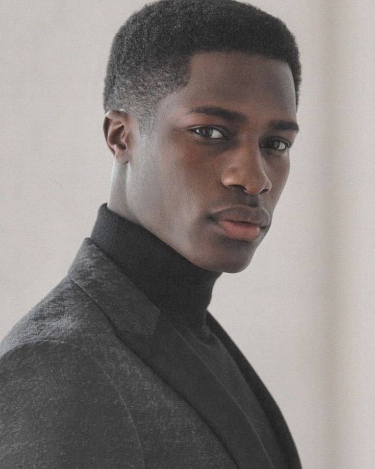 a closeup portrait of a handsome, darkskinned man with short, neatly styled hair. he is wearing a black turtleneck sweater and a textured gray suit jacket. he is looking directly at the camera with a serious expression. the shot is welllit, highlighting his sharp jawline and clean shave. the background is a neutral color, focusing attention on the subject.