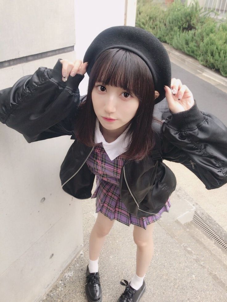 a young asian woman in a trendy outfit stands in front of a concrete wall and looks at the camera.  she is wearing a black beret, black leather jacket, purple plaid skirt, white shirt, white socks, and black shoes.  she has her black hair in bangs and is standing with her left leg slightly bent.  she is standing in a sunny area with a paved sidewalk in front of her.