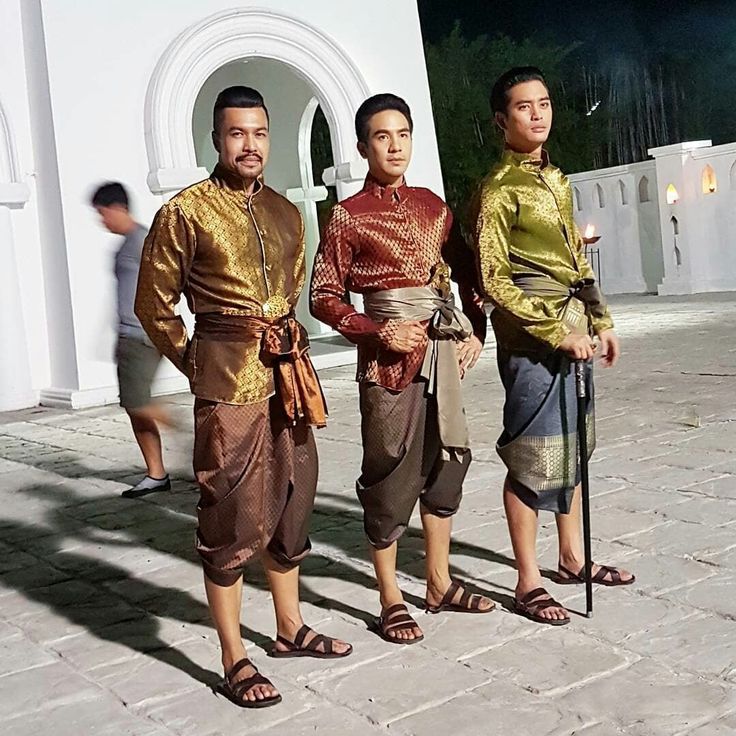 three men wearing traditional thai clothing, standing outdoors on a stone paved ground. they all appear to be smiling as they look toward the camera.