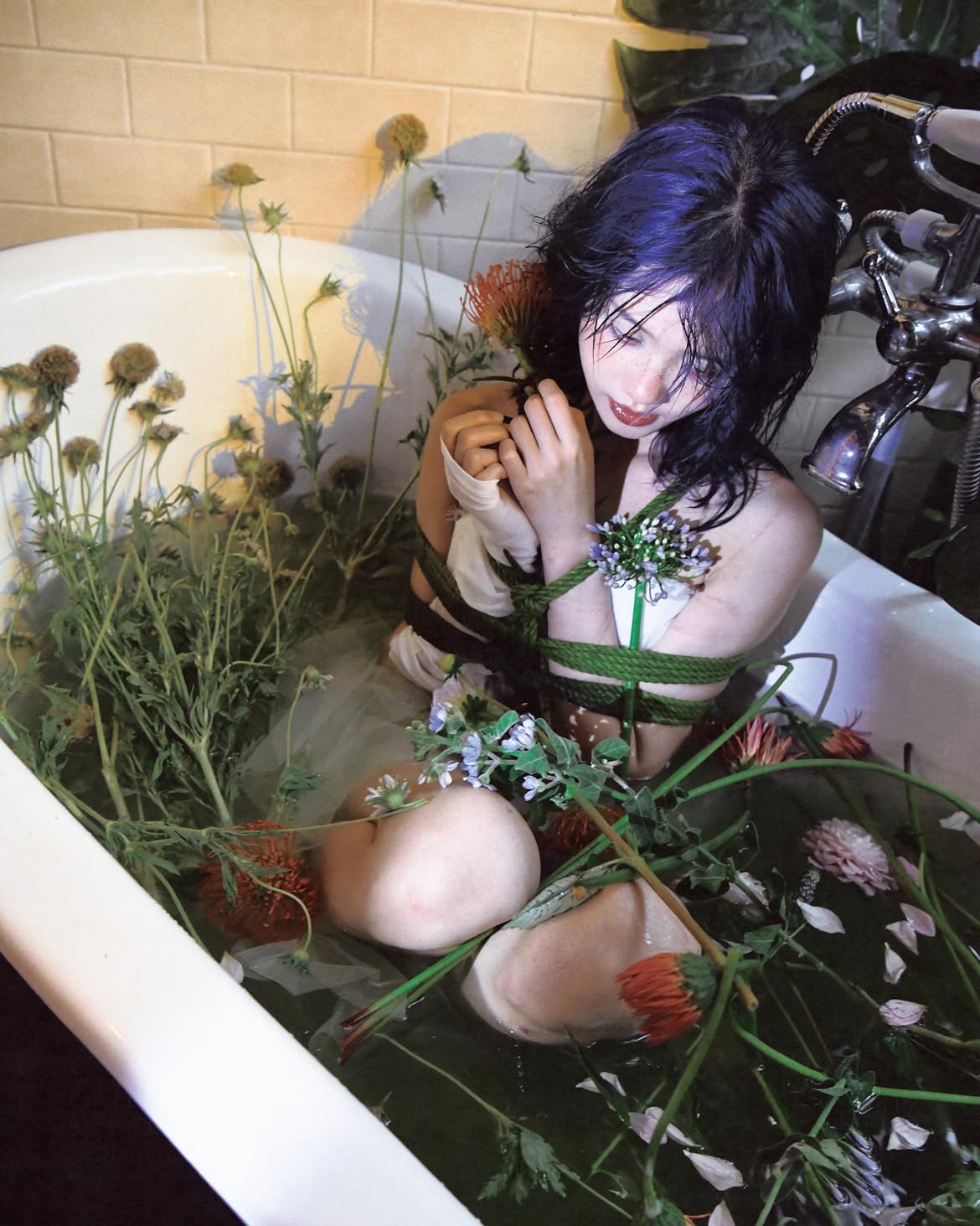 a pale woman with purple hair is in a white bathtub filled with water, plants, and flowers. her hands are tied with rope, and she appears to be in a state of captivity or bondage. the bathtub has a classic faucet and is set against a tiled wall.