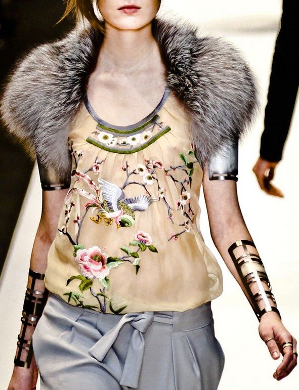 a fashion model is wearing a fur collar, a delicate floral blouse, silver bracelets on her arms, and grey pants. her makeup is natural, and her hair is swept away from her face. she is looking off to the side, and her pose is relaxed and confident.