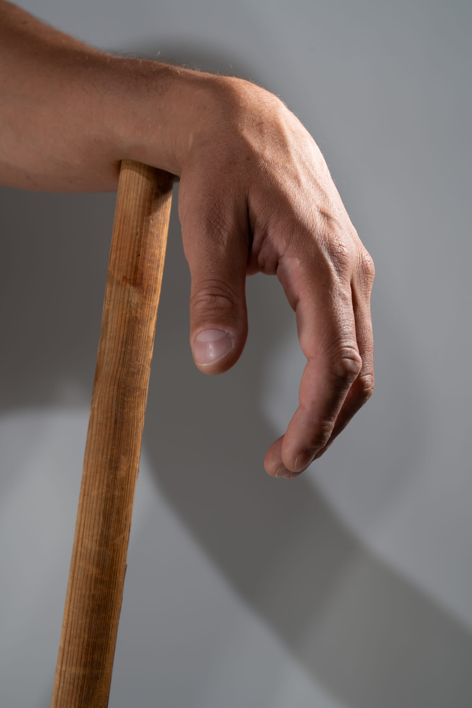 a closeup image of a man's hand gripping a wooden stick against a light gray background. the hand is illuminated by studio lighting, creating a strong shadow on the wall.
