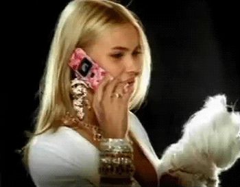the image shows a blonde woman with long hair, wearing a white top and substantial gold jewelry (necklace and bracelets). she is holding a small, pink flip phone to her ear and appears to be talking on it.  a small, fluffy white dog, possibly a maltese, is partially visible in the lower right corner, seemingly being held or near the woman. the overall aesthetic is reminiscent of early 2000s fashion and styling. the lighting and background suggest a professional photoshoot or video shoot.