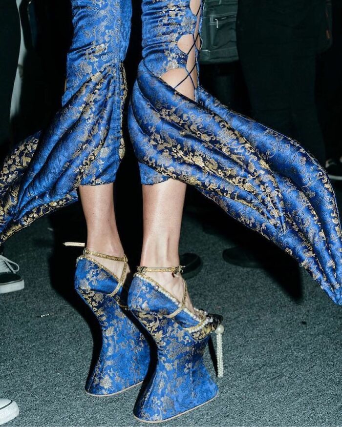 a person wearing blue and gold patterned clothing and shoes. the pants have large, padded sections that flare out from the lower legs. the shoes are matching high heels with a platform and ankle straps, also adorned with pearls. the style appears to be avantgarde fashion.