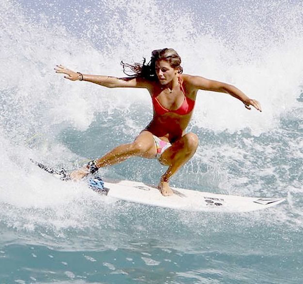 a woman in a bikini surfs a wave on a surfboard. she is midair, with her arms outstretched and her legs bent. the water is splashing around her, and the sun is shining.