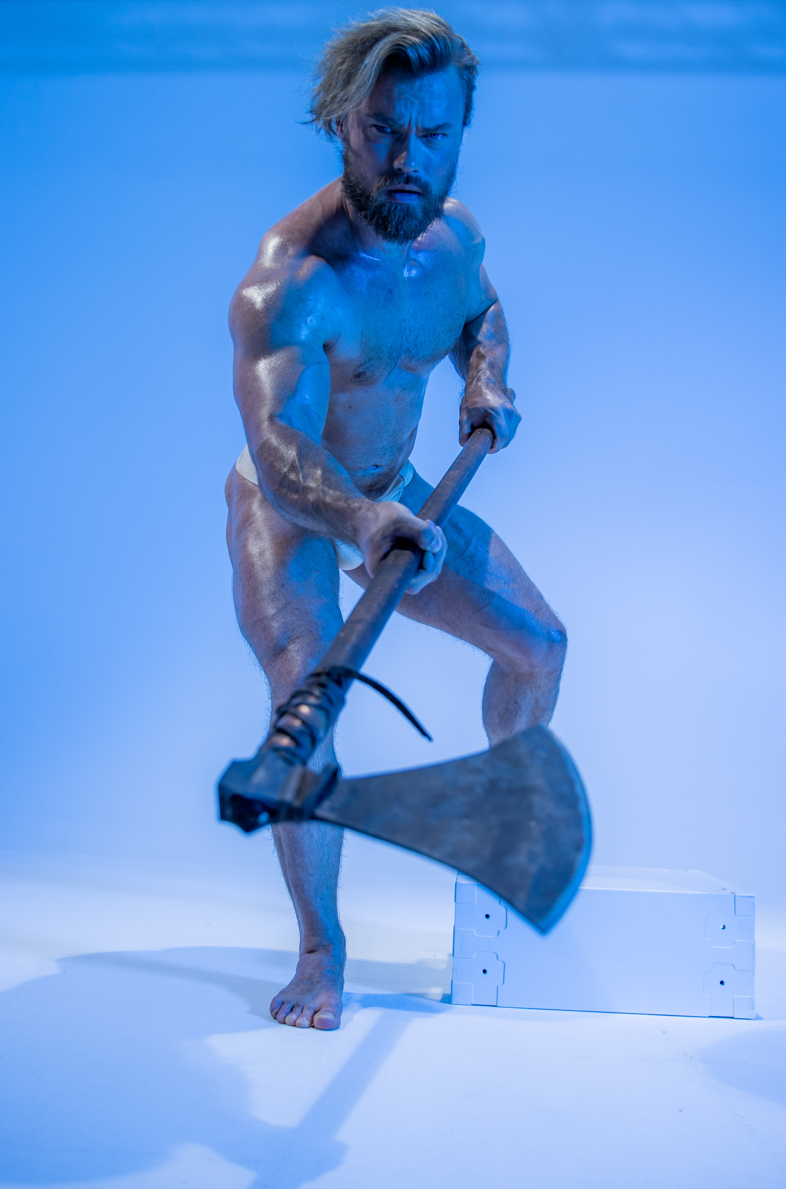 a muscular man with long blond hair and a beard stands in a fighting pose against a bright blue backdrop. he is only wearing a beige speedo and wields a large, doubleheaded axe. his body glistens as if wet and he glares directly at the viewer with an intense expression. there is a white box on the ground behind and to the right of the man.