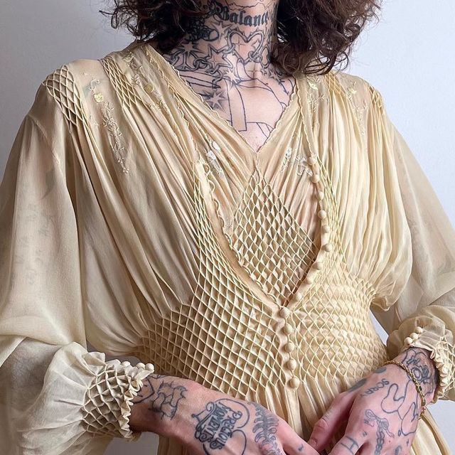 a person is wearing a beige vintage chiffon dress with long sleeves and ruffled cuffs. the dress has smocking detail on the bodice and buttons down the front. the person has tattoos on their neck, chest, and hands. the dress has a vneck. the background is a plain white wall.