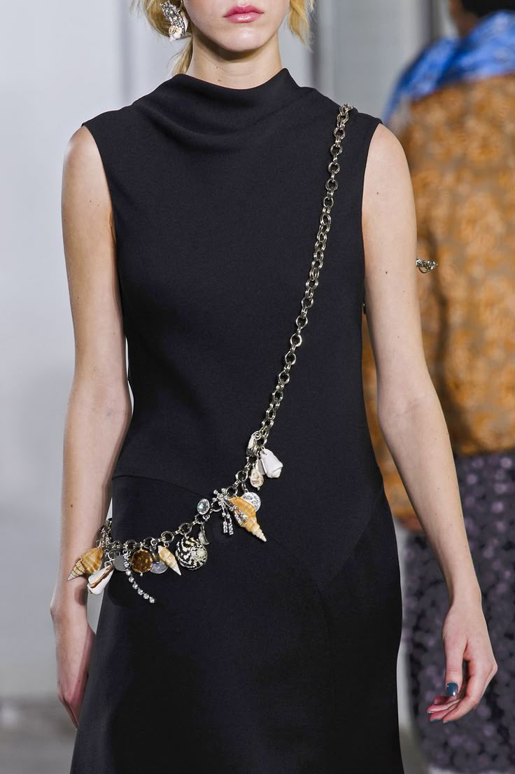 a runway model wearing a black sleeveless high neck dress with a chain belt embellished with seashell charms. she is also wearing earrings. the background is blurry with another model partially visible.