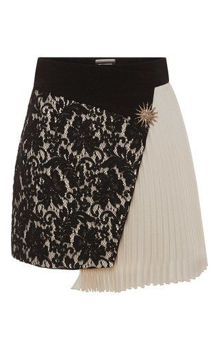 a closeup shot of a stylish asymmetrical skirt. the skirt has a black waistband, with the left side covered in black lace with a cream background. the right side features pleated creamcolored fabric. a gold sunburst brooch is placed on the front where the two fabrics meet.