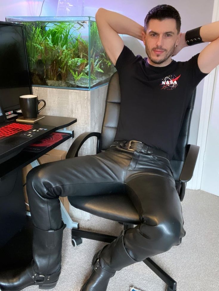 a man is sitting in an office chair. he is wearing black leather pants and boots and a black nasa tshirt. his arms are behind his head in a relaxed pose. he is sitting in front of a computer and a fish tank with plants. the overall setting appears to be a home office, casual but stylish.