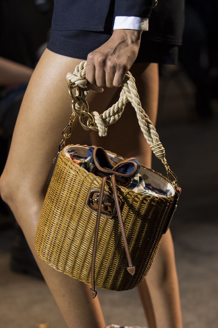 a closeup shot of a model holding a wicker handbag with a rope handle and gold hardware. the bag features a drawstring closure and is displayed at a fashion show.