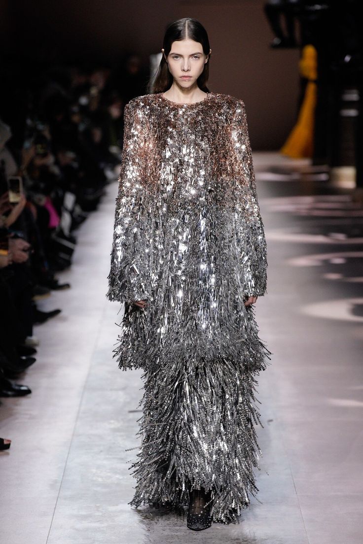 a female model is walking down a runway wearing a floorlength, longsleeved dress covered in metallic silver fringe that transitions into a brown hue at the top. the dress is shimmery and textured, resembling sequins and fringe. the runway show features an audience on the left and stage lighting in the background.