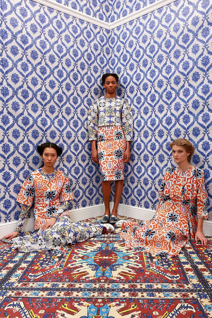 hree women models are sitting on a vibrant patterned rug in a room with matching blue and white patterned wallpaper. the women are wearing similar patterned dresses and skirts in shades of blue and orange, creating a cohesive and visually striking image. the room's corner and the intricate patterns on the wall and floor are emphasized in the composition.