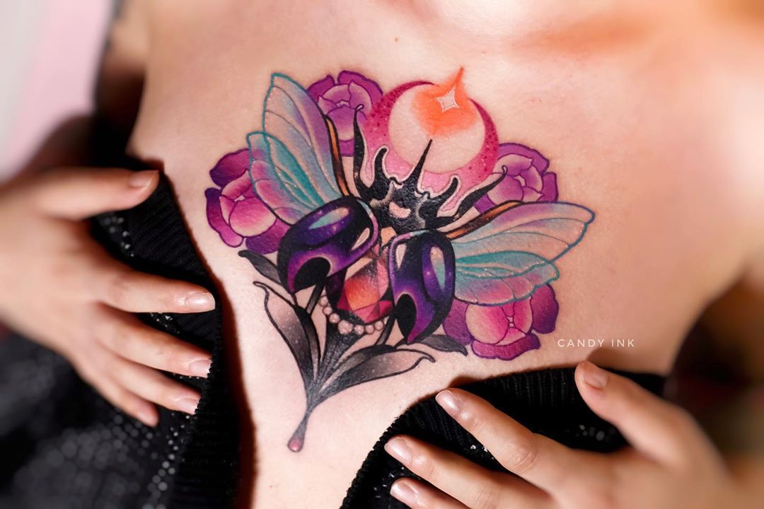 a closeup photograph showcasing a vibrant neotraditional tattoo of a jeweltoned beetle with delicate, iridescent wings, nestled amongst a cluster of pink flowers, inked on a woman's chest.