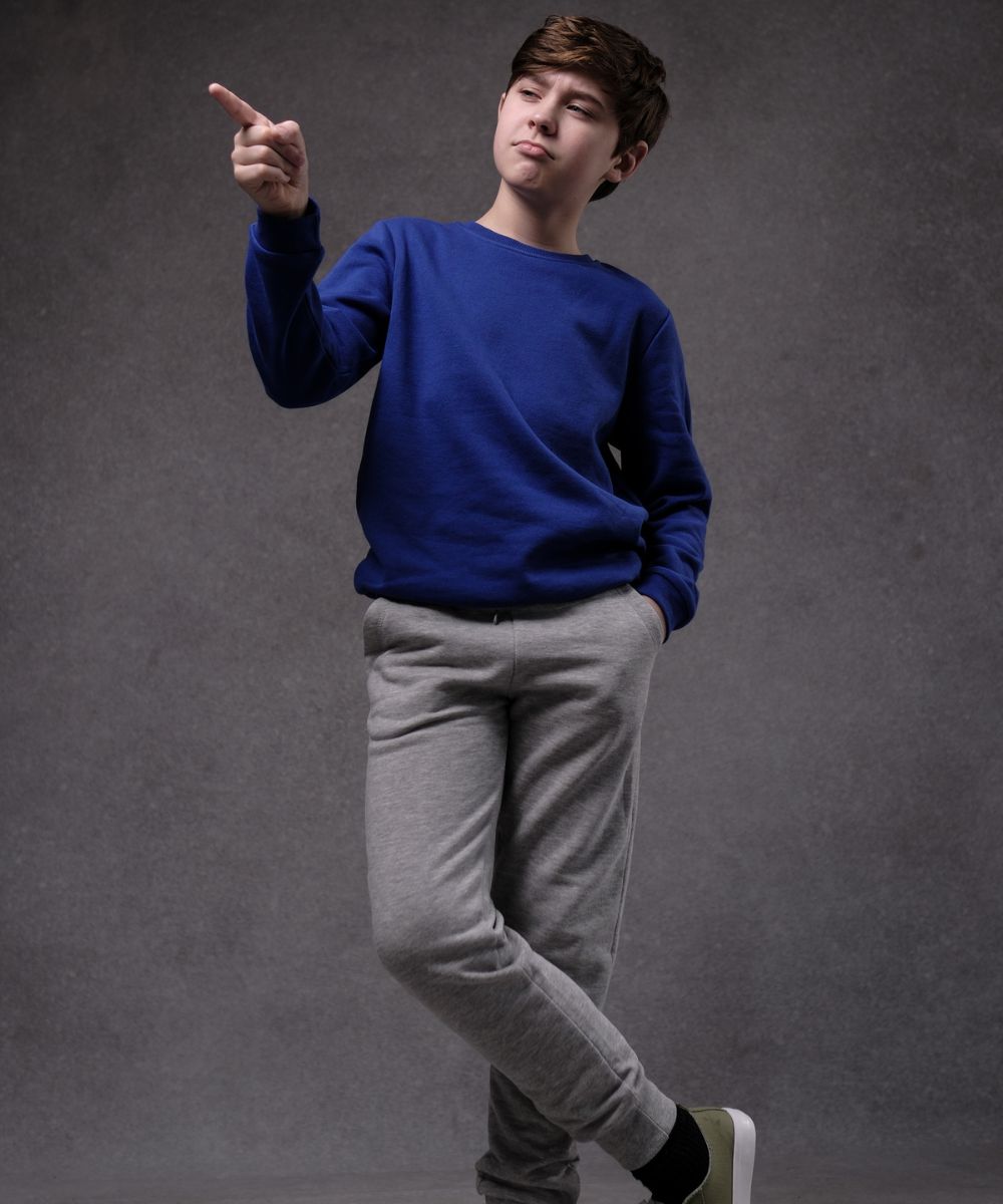 a full shot of a teenage boy standing against a grey background. he's wearing a royal blue sweatshirt and grey sweatpants.  his right hand is pointing to the left. he has a serious or slightly skeptical expression on his face.  he is angled slightly and his pose is relaxed but also somewhat assertive.  the lighting is even, creating a clear and welllit image.