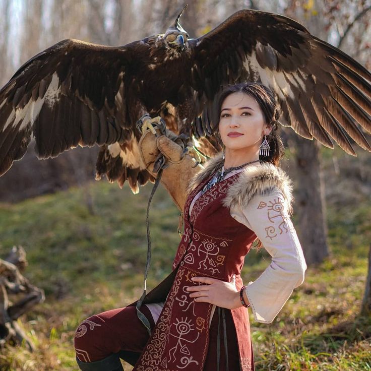 a young woman in traditional clothing, with a furtrimmed vest, stands in the forest, holding a golden eagle on her outstretched arm. the eagle's wings are spread wide behind her, and its head is turned to look at the camera. the woman is looking directly at the camera, with a serious expression on her face. the image is taken outdoors in a natural setting, and the background is a blurry mix of trees and foliage. the woman and the eagle are the focus of the image. the eagle is a powerful symbol in many cultures, and this image captures the beauty and majesty of this bird.  the woman's traditional dress is a beautiful example of cultural heritage. the image is a stunning portrait of a woman and her eagle.