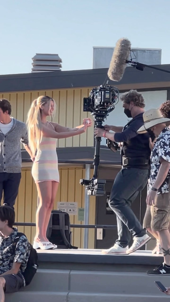 a woman in a striped dress stands on a platform surrounded by a film crew. a large camera and boom microphone are visible. the woman is gesturing with her hands. other people are on and around the platform.