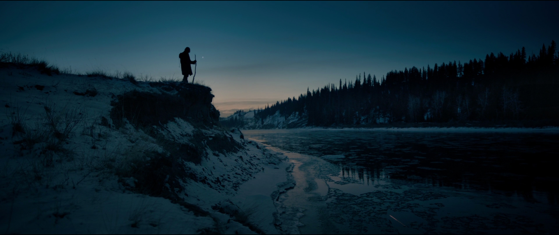 a lone man stands silhouetted against the twilight sky on a snowy riverbank, his spear held upright. a partially frozen river, reflecting the fading light, stretches out before him, leading to a dark forest on the opposite bank.  the overall palette is dark and cold, emphasizing the harsh beauty and isolation of the scene.