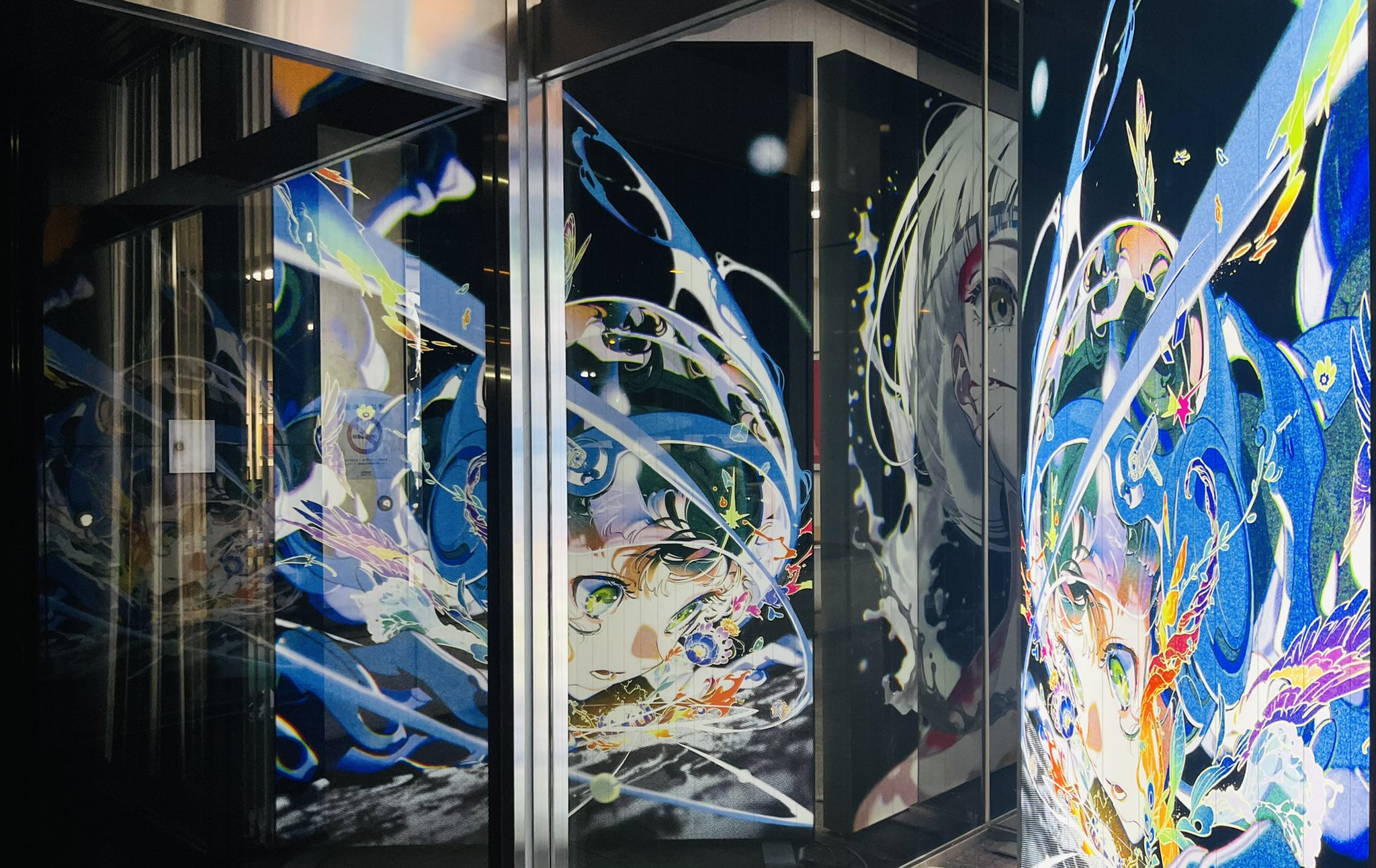 the image showcases a vibrant, digitallystyled artwork displayed on the glass facade of a building, likely at night. the artwork features animestyle characters amidst abstract swirls of blue, teal, and white, creating a sense of dynamic energy. reflections of the city lights mingle with the artwork, adding another layer of visual complexity.