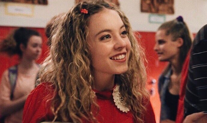 the image is a closeup shot of a young woman, possibly a teenager, with shoulderlength, curly light brown hair. she's wearing a red dress with a white pearl collar and has small red hair clips in her hair. she has a cheerful expression and is smiling. the background is slightly out of focus but appears to be a school setting, with red lockers and other students visible. the overall impression is a cheerful and casual portrait, possibly a still from a film or television show.