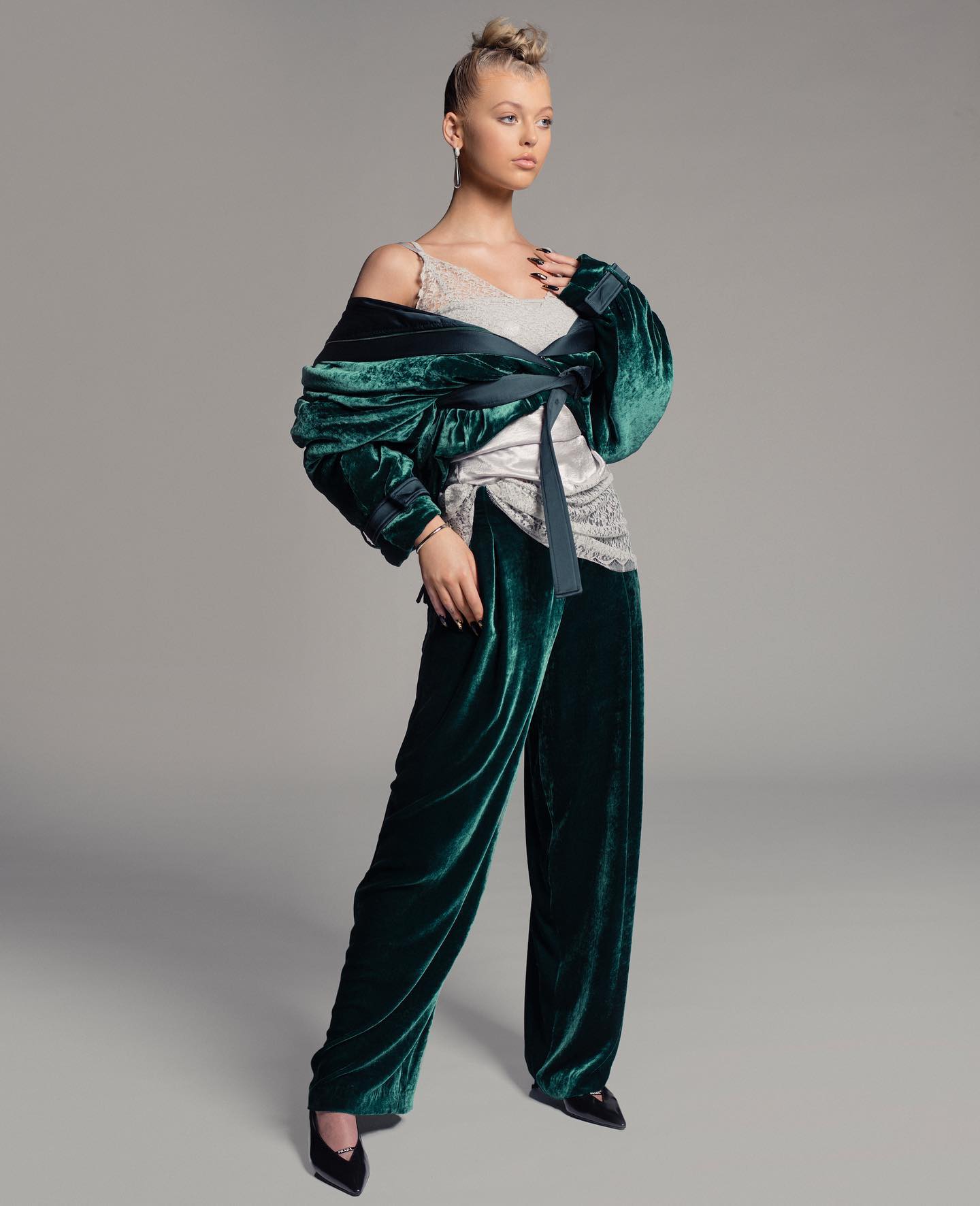 a fulllength portrait of a woman wearing a stylish, green velvet outfit. she is positioned against a neutral gray background. the outfit consists of widelegged velvet pants, a lacetrimmed blouse, and a matching velvet wrap tied at the waist with a dark green ribbon. the woman has her hair styled in a high bun and is wearing delicate jewelry. she is looking off to the side with a confident expression. the overall aesthetic is sophisticated and elegant.