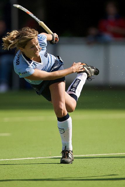 a female field hockey player takes a shot on goal. the player is in mid air and has her stick raised to strike the ball. the player is wearing a blue and white jersey and black shorts. the player is wearing white socks and black cleats. the player is playing on a green field. the player is surrounded by other players, but they are not in focus.