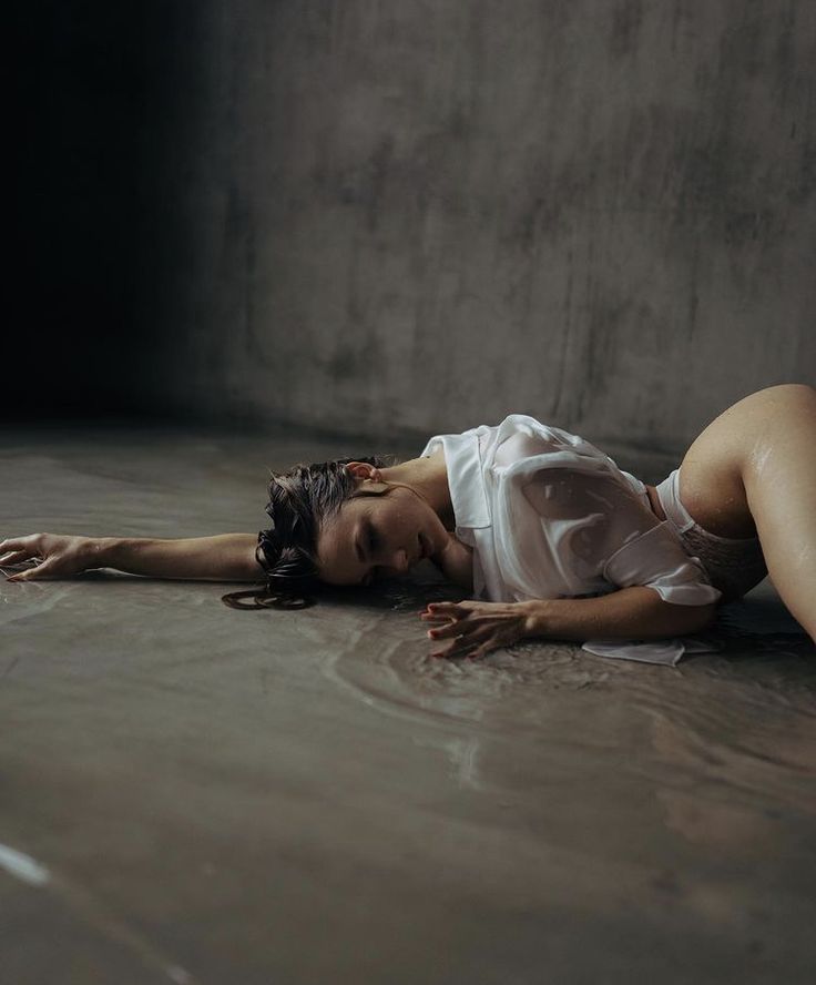 a woman lies on a wet concrete floor, her body slightly angled, in a white, wet shirt. her hair is wet and spread around her head. she appears to be sad or vulnerable, her eyes closed and her expression serene yet heavy. the setting is dark and moody, enhancing the emotional impact of the image. the water on the floor reflects light subtly, giving it a reflective quality. the overall mood of the photo is somber and sensual.