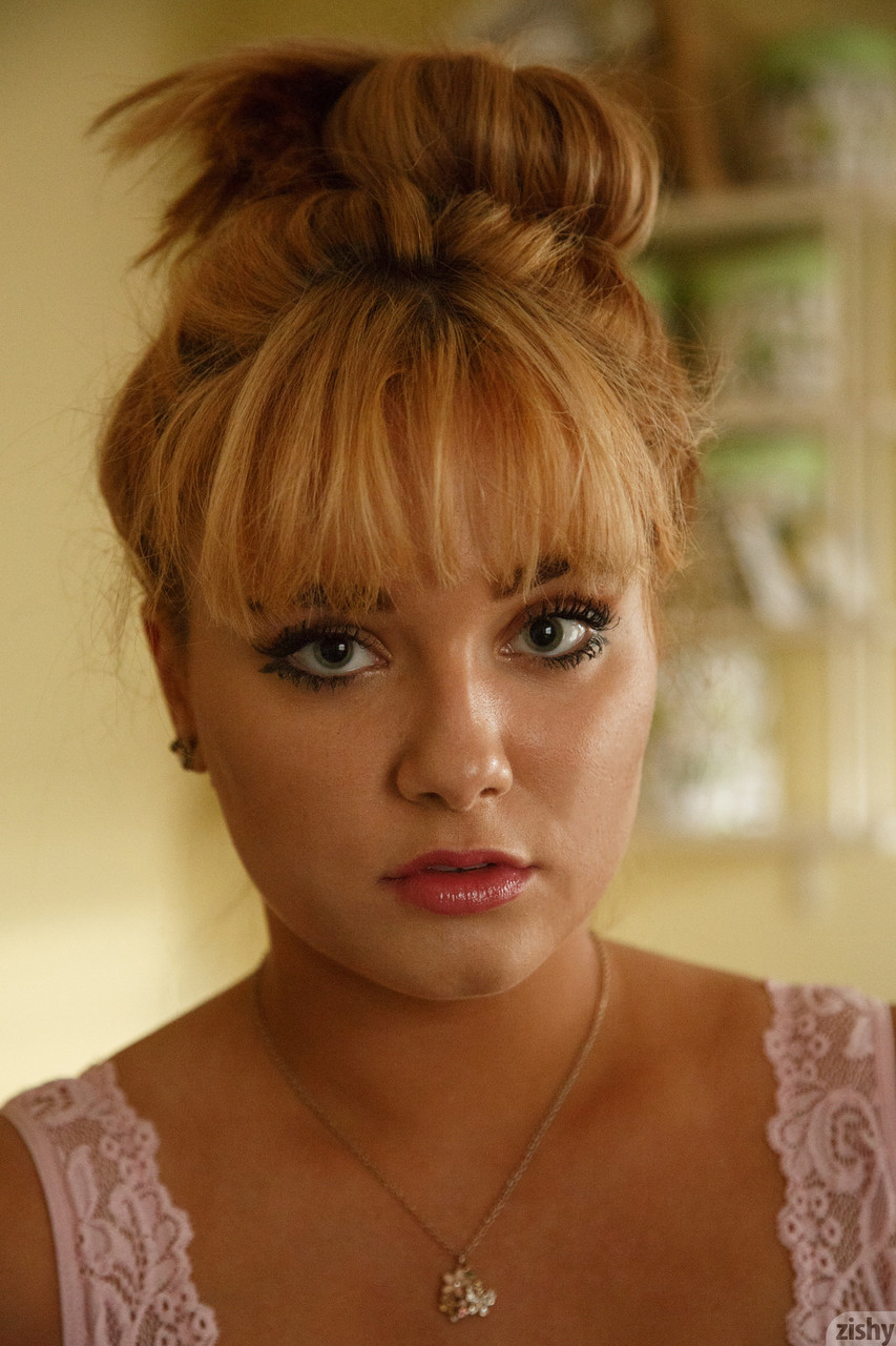 a young woman with blonde hair styled in a high bun poses for a portrait. she is wearing a delicate, pale pink lace top and a silver necklace with a small pendant. her makeup is subtle, with a focus on her eyes and lips. the background is blurred, suggesting a home environment, and the lighting is soft and natural.