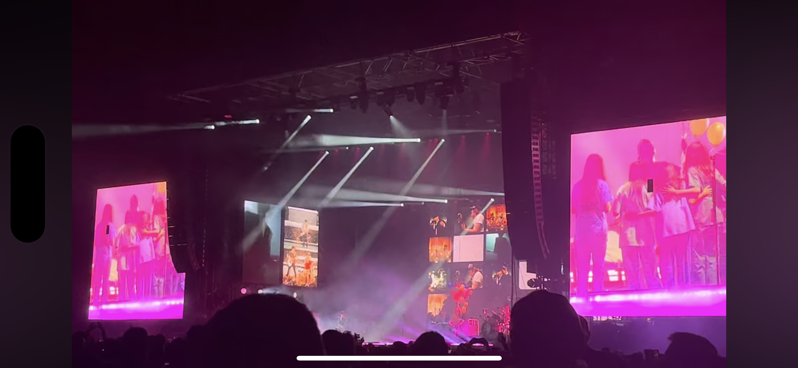 a concert stage is lit up with bright spotlights, illuminating a large crowd gathered below. two large video screens on either side of the stage showcase a band performing, their images bright and captivating against the dark background. the energy is palpable, suggesting a lively and exciting atmosphere. the crowd's enthusiasm for the performance is evident, creating a vibrant spectacle of music and entertainment.