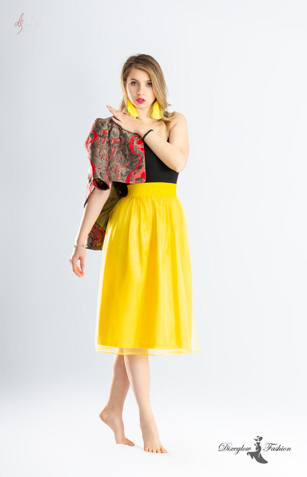 a full shot of a beautiful blonde woman model wearing a bright yellow midi skirt, a black top, and a red and grey patterned jacket. she is wearing yellow earrings and is barefoot.  the woman is posing against a white background in a studio setting. the image is stylish and elegant, showcasing the clothing.  watermarks for the photographer and fashion brand are visible.