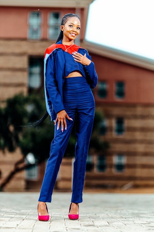 a smiling young african woman, possibly a recent graduate, poses confidently for a portrait. she's wearing a vibrant blue pantsuit, styled with a shorter cropped jacket and highwaisted trousers. the suit is complemented by a colourful, capelike graduation stole, partially visible, with hints of red and light blue.  she's wearing bright pink high heels and has her long dark hair in neat braids. the background is an outoffocus urban setting with a red brick building visible.  the overall impression is one of success, achievement, and stylish professionalism.