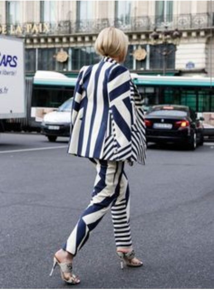 a stylish blonde woman wearing a black and white striped blazer and matching pants walks down a parisian street.  she's wearing highheeled mules and has a bob haircut. the background features buildings and a street scene. the overall image is one of high fashion and street style.