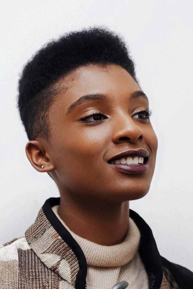 a closeup portrait of a young black woman with short hair. she is smiling and looking up, her eyes are brown and her lips are painted with a dark brown lipstick. she is wearing a brown jacket with a beige turtleneck sweater underneath. the background is a white wall.