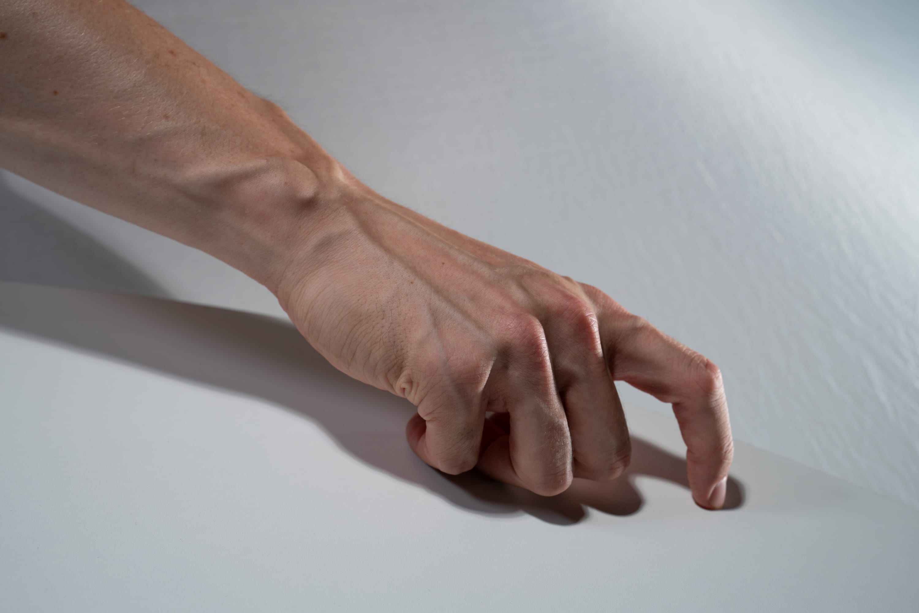 a closeup photo of a fair skinned person's left hand with pronounced veins.  the fingers are curled in and the hand is resting on a white surface. the lighting is soft with a light source from above.  there is a subtle shadow from the hand falling to the left on the white surface.