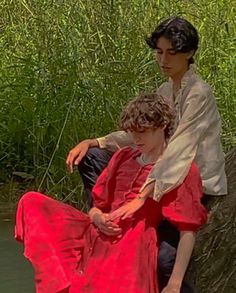 a couple in periodstyle clothing are seated near a creek. the person in the red dress is resting their head on the shoulder of the person in the white shirt. the person in the white shirt has their arm around the other person's waist and is holding their hand. they are both looking down. the background is lush greenery.