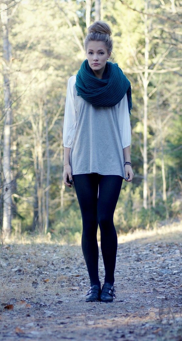 a young woman with blonde hair in a bun, wearing a teal scarf, gray shirt, and black leggings and boots, is standing in a forest setting. she is looking directly at the camera.